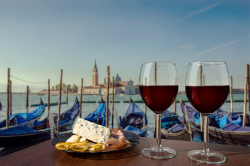 Two glasses of wine with charcuterie assortment on view of gondolas and San Giorgio Maggiore church in the background in Venice. Glass of red wine with different snacks - plate with ham, sliced