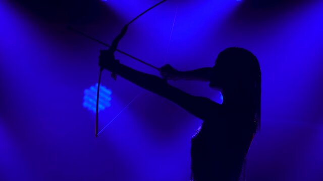 Silhouette of dancing Joan of Arc with bow and arrow on stage in a dark studio with smoke and neon lighting. A bright young woman in a gold dress and a headdress. Dynamic neon lighting effects