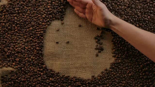 Appearance of coffee cup icon made of beans on jute cloth by slow motion waving of woman's hand. Coffee piktogramm designed. Natural, authentic coffee traditions.