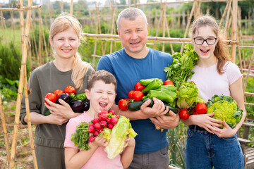 Portrait of happy parental couple with teen daughter and preteen son posing with fresh harvest in backyard garden.