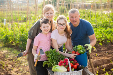 Portrait of happy friendly family with two teen children standing with crop of vegetables in garden on sunny summer day.