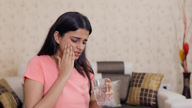 Portrait of a beautiful Indian girl with a severe toothache and dental problems. Attractive young female feeling pain in her tooth while drinking a glass of cold water - tooth sensitivity concept