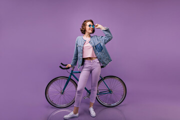 Slim blithesome woman posing in studio with bicycle. Indoor full-length shot of curly female model in purple pants.