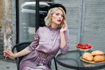 A girl in a dress sits at a table with champagne and French rolls. Bright red lipstick and shoes. © Denys