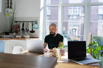 Freelancer programmer working from home on kitchen and using laptop. Bearded man working with a computer, prints text on keyboard. Self entrepreneur sitting and working at his modern apartment.
