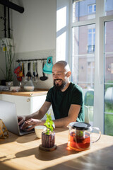 Freelancer programmer working from home on kitchen and using laptop. Bearded man working with a computer, prints text on keyboard. Self entrepreneur sitting and working at his modern apartment.