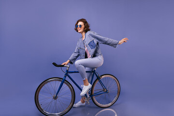 Amazing woman in spring clothes sitting on bicycle. Indoor portrait of lovely girl in sunglasses fooling around on violet background.