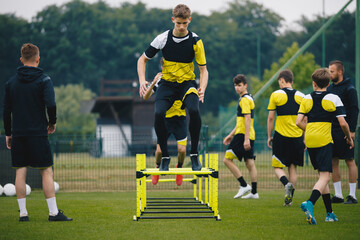 Soccer training drills with hurdles agility. Football players jumping high over hurdles obstacles. Footballers in a team on practice pitch. Two coaches coaching junior level teenage players