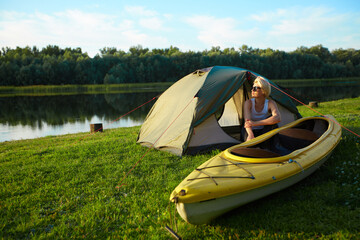 Travel, kayaking and hiking concept. Portrait of young beautiful woman sitting near green tent with kayak near river.