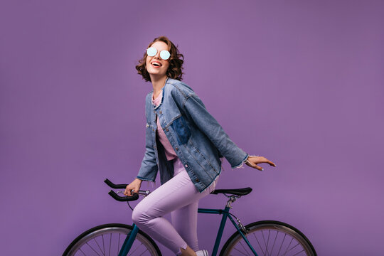 Glamorous female model in sparkle glasses posing with bicycle. Pretty curly girl expressing positive emotions on purple background.