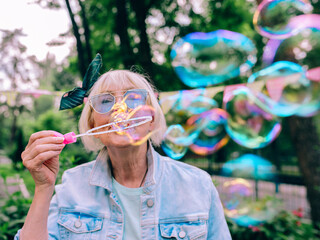 senior (old) stylish woman with gray hair and in blue glasses and jeans jacket blowing bubbles...