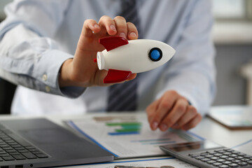 Close up of male hand holding plastic rocket while sitting at desk with laptop and financial papers