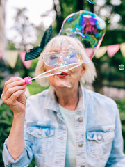 senior (old) stylish woman with gray hair and in blue glasses and jeans jacket blowing bubbles outdoors. Holidays, party, anti age, fun concept
