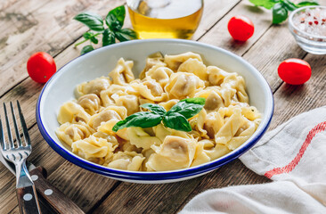 Tortellini with cheese sauce, basil leaves in bowl on rustic wooden table.