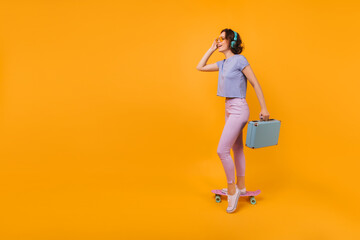 Shapely lady in white gumshoes posing with blue suitcase in studio. Photo of good-humoured short-haired woman standing on longboard.