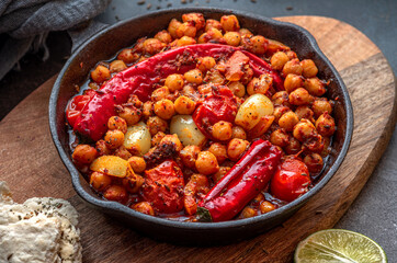 Spicy chickpeas with hot red peppers, tomatoes, cumin, garlic and other spices in a frying pan on a black background. Chickpeas stewed with vegetables and spices, oriental cuisine. Vegetarian food.
