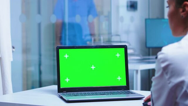 Over shoulder footage of doctor using laptop with copy space available in hospital . Nurse in uniform arriving in cabinet. Medic wearing uniform using notebook with chroma key on display in medical