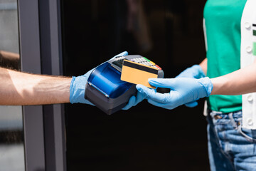 Cropped view of woman in latex gloves holding credit card near courier with payment terminal on urban street