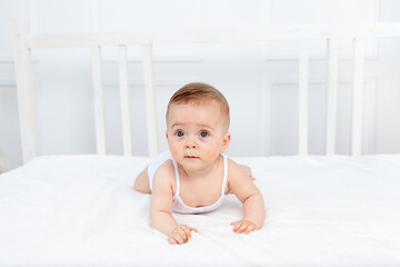 Fototapeta premium smiling baby girl 6 months old lies on the bed in the nursery on her stomach and looks away, morning baby, baby products concept