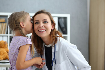 Cheerful woman doctor with stethoscope is listening to kid patient whispering to her ear in office