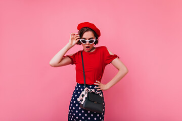 Unpleased girl with black handbag posing in french beret. Studio portrait of slim woman in red attire isolated on pink background.