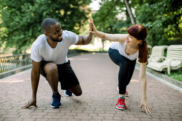 Young fitness couple of African bearded man and attractive Caucasian woman, ready to start running at the park, giving high five each other and smiling. Jogging together outdoors