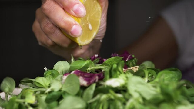 Woman's hand squeezes lemon onto green healthy salad of endive, corn salad and baby spinach.