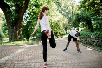 Young multiethnic jogging couple, Caucasian red haired girl and African guy, warming up and stretching before morning running in the city park