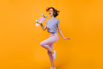 Fototapeta na wymiar Full-length portrait of happy caucasian female model with pink longboard. Studio photo of beautiful curly girl listening music in headphones on orange background.