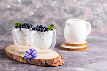 Chia pudding with mint and blueberries in glasses on a wooden stand with a blue flower and cream jug on a light background