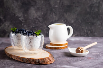 Chia pudding with mint and blueberries in glasses on a wooden stand with a honey stick and cream jug on a dark background