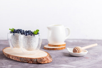Chia pudding with mint and blueberries in glasses on a wooden stand with a honey stick and cream jug on a light background
