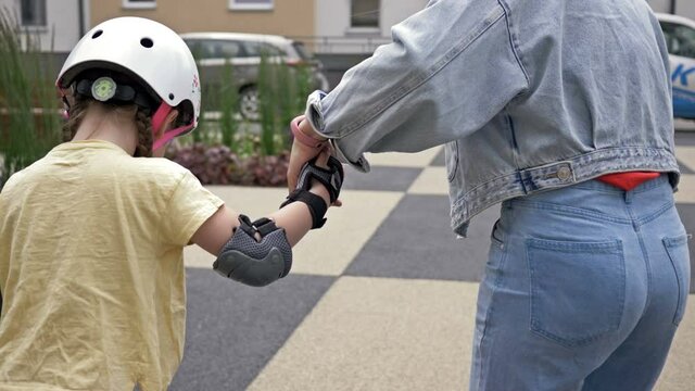 Mom teaches little daughter to roller skate. The woman holds the child by the hand. Back view.