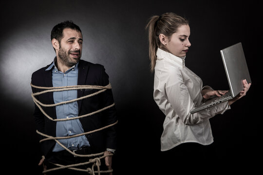 uomo moro in giacca scura e camicia blu, legato con una corda, &egrave; disperato vicino alla dsua compagna bionda che guarda un computer portatile stando in piedi vicino a lui,  isolati su sfondo nero
