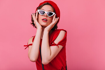 Glamorous french lady touching her sunglasses during indoor photoshoot. Studio shot of magnificent white woman with short wavy hair.
