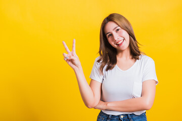 Asian Thai happy portrait beautiful cute young woman smile standing wear t-shirt showing finger making v-sign symbol near eye looking to camera, isolated studio shot yellow background with copy space