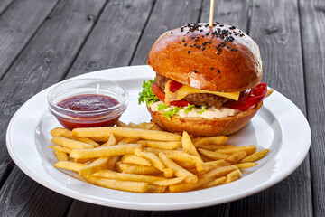 Hamburger with chicken cutlet, cheese, french fries, tomato, salad, sauce and ketchup on a white plate and a dark wooden background