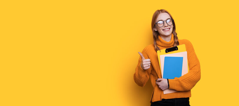 Lovely red haired student with freckles gesturing the like sign and holding folders on a yellow wall with free space