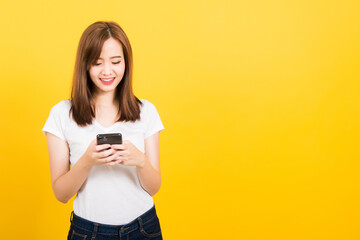 Fototapeta premium Asian happy portrait beautiful cute young woman teen smile standing playing game or writing message on smartphone looking to the phone isolated, studio shot on yellow background with copy space