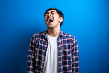 Fototapeta premium Positive cheerful male closing eyes tight cannot stop laughing at good joke. Handsome young man of Asian appearance with black hair wearing flannel shirt stands on blue background in Studio