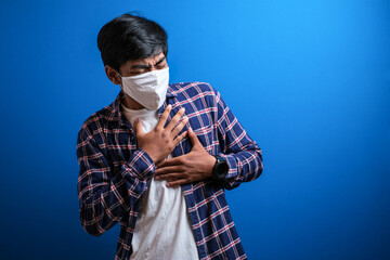 Asian young student wearing flannel shirt feel pain on his chest and wear protective mask to prevent the spread of corona virus against blue background