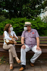 Photo of a middle age couple sitting on a bench taking a selfie and wearing white face masks in the park	
