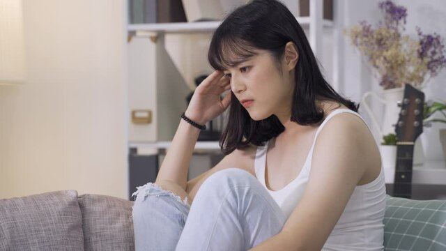 asian girl curling up on sofa is in anguish over her quarrel with boyfriend. vexed looking  chinese young woman having no idea how to fix relationship with lover, touching face and hair with anxiety.