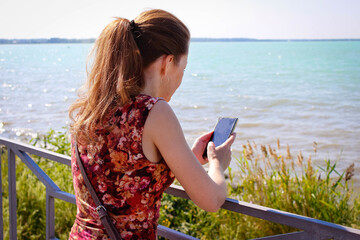 A girl with red hair stands on the embankment with a smartphone in her hands.