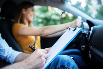 Close up view of driving instructor holding checklist while in background female student steering and driving car. Acquiring driver's license. - obrazy, fototapety, plakaty