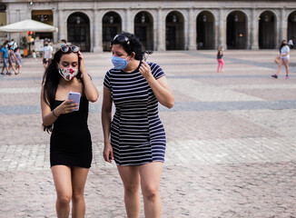 Young women walking around a square in the city using medical face masks while using their electronic devices