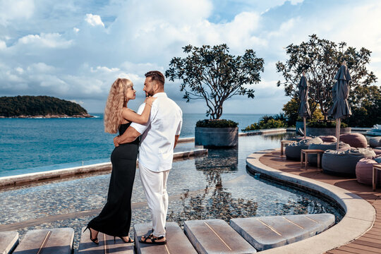 A romantic couple in love stands near the infinity pool in a luxury villa. A man in a white suit hugs his woman in a black dress. - Powered by Adobe