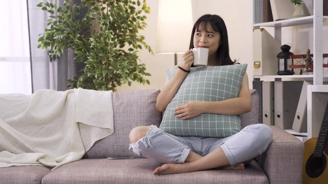 lovely asian lady having tea in a relaxing mood on living room couch. Young lady in love holding cushion beamed with happiness thinking of her thoughtful boyfriend.