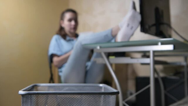 girl imposingly sits at a table and throws crumpled pieces of paper in a trash bin. Selective focus on bin