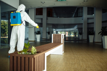 A man wearing protective suit is disinfecting a bench in an empty shopping mall with sanitizing spray. Cleaning up the public place to prevent covid spread. Healthcare precautions and safety concept.